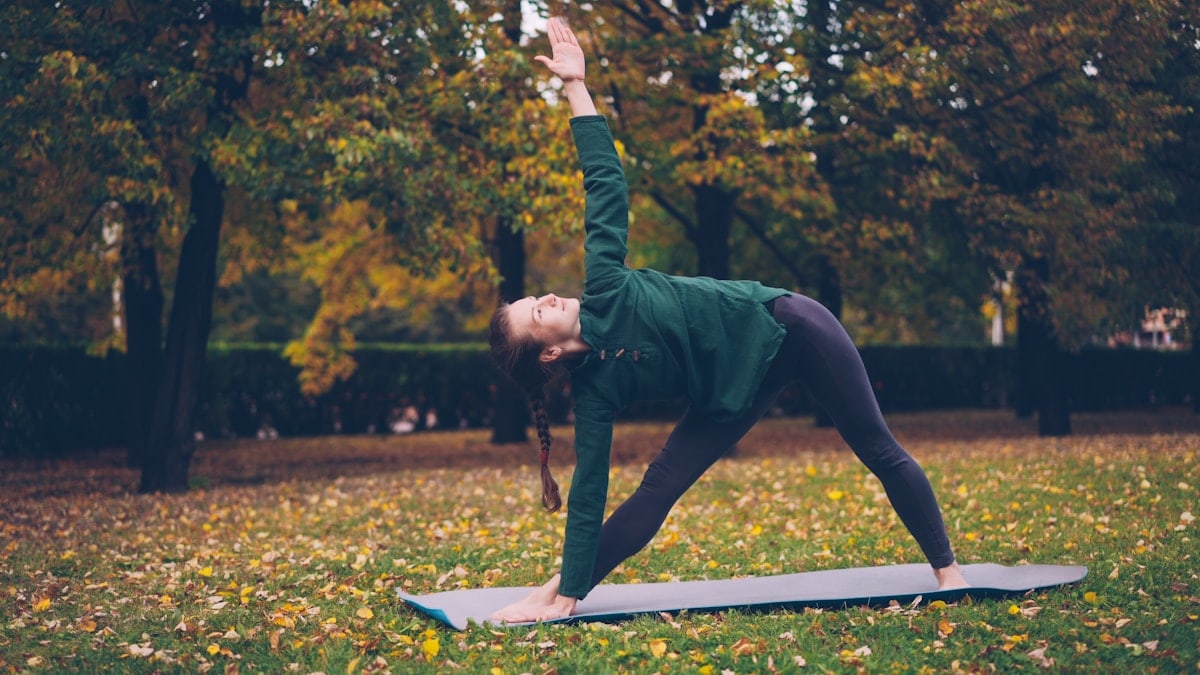 Cheerful young woman is doing yoga balancing exercises during individual practice in city park standing on mat and enjoying rest and nature. People, healthy lifestyle and sports concept.